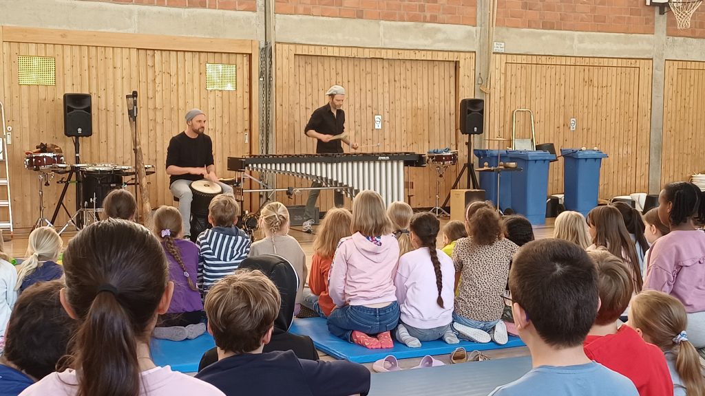 zwei Männer musizieren vor Kindern in der Turnhalle